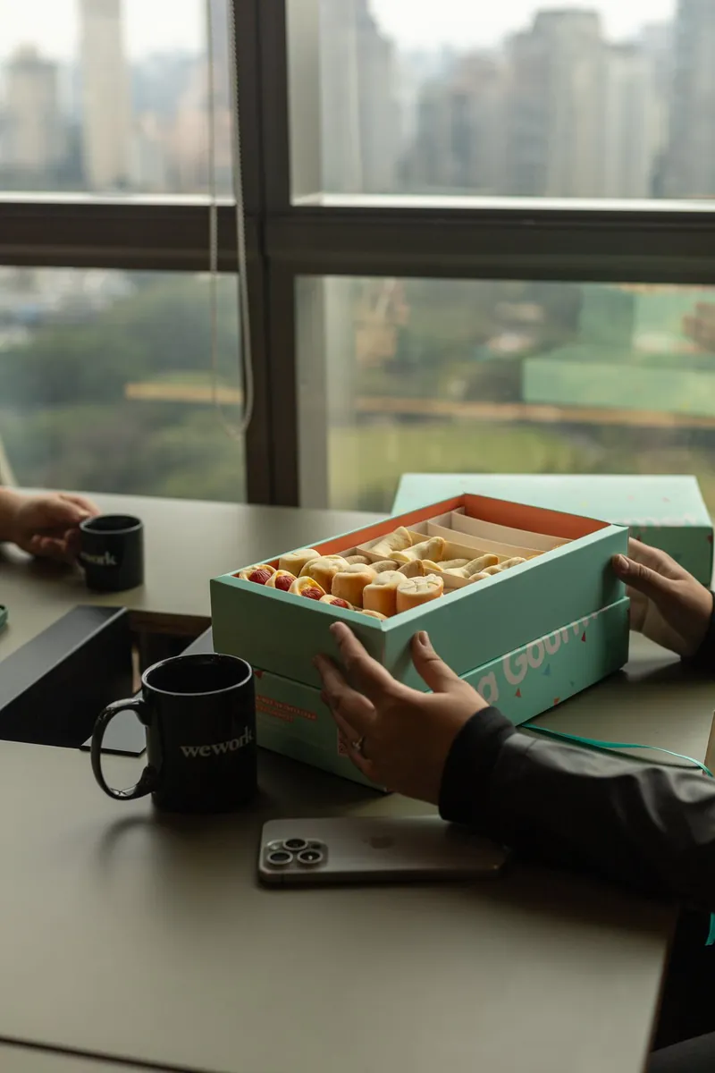 Caixa de coffee break corporativo Caixa Gourmet aberta sobre mesa de reunião com vista para o skyline de São Paulo ao fundo.